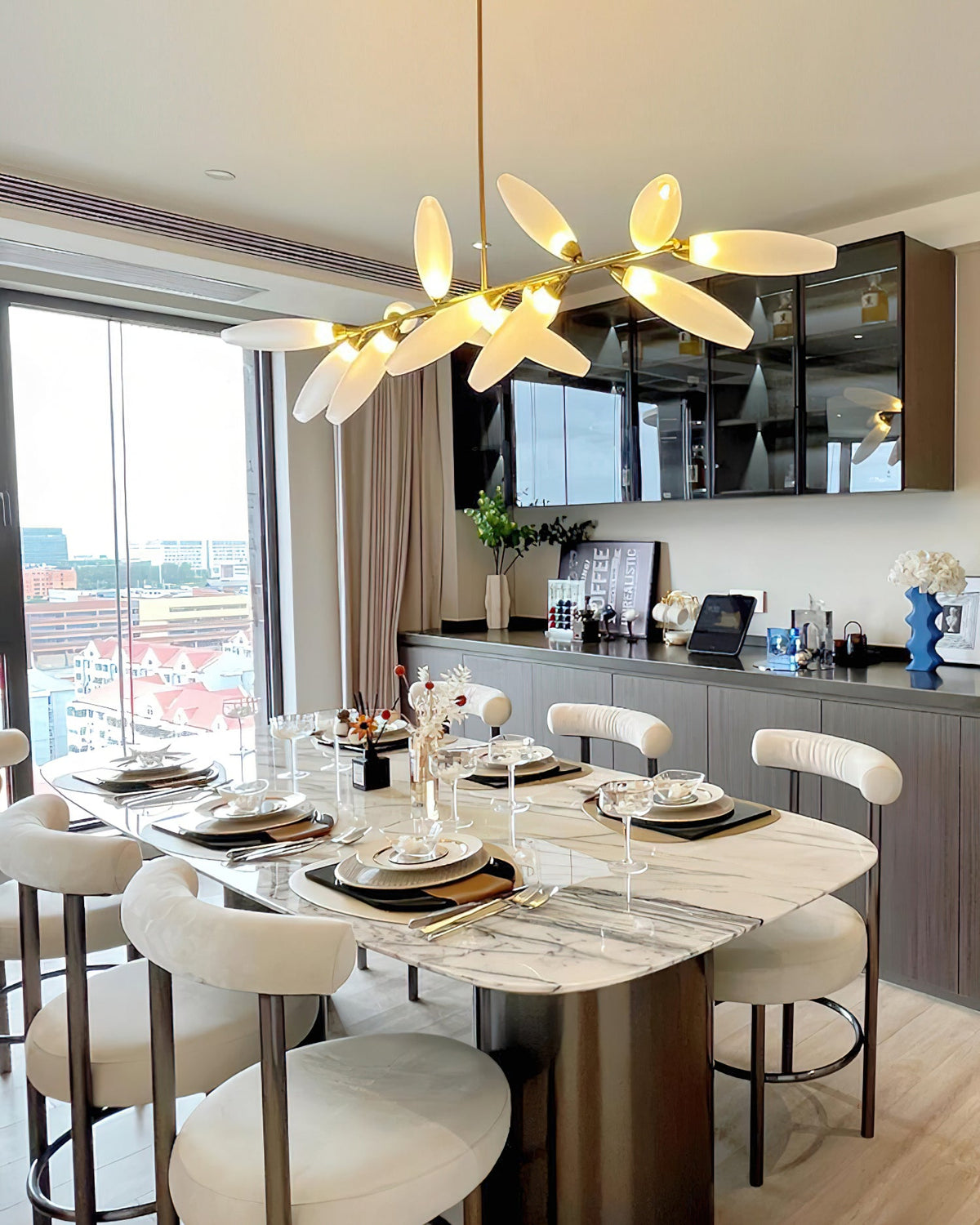 A wider view of a Clérachic chandelier lit warm above a marble dining table and cream bouclé chairs, with a glazed cabinet and a city view.