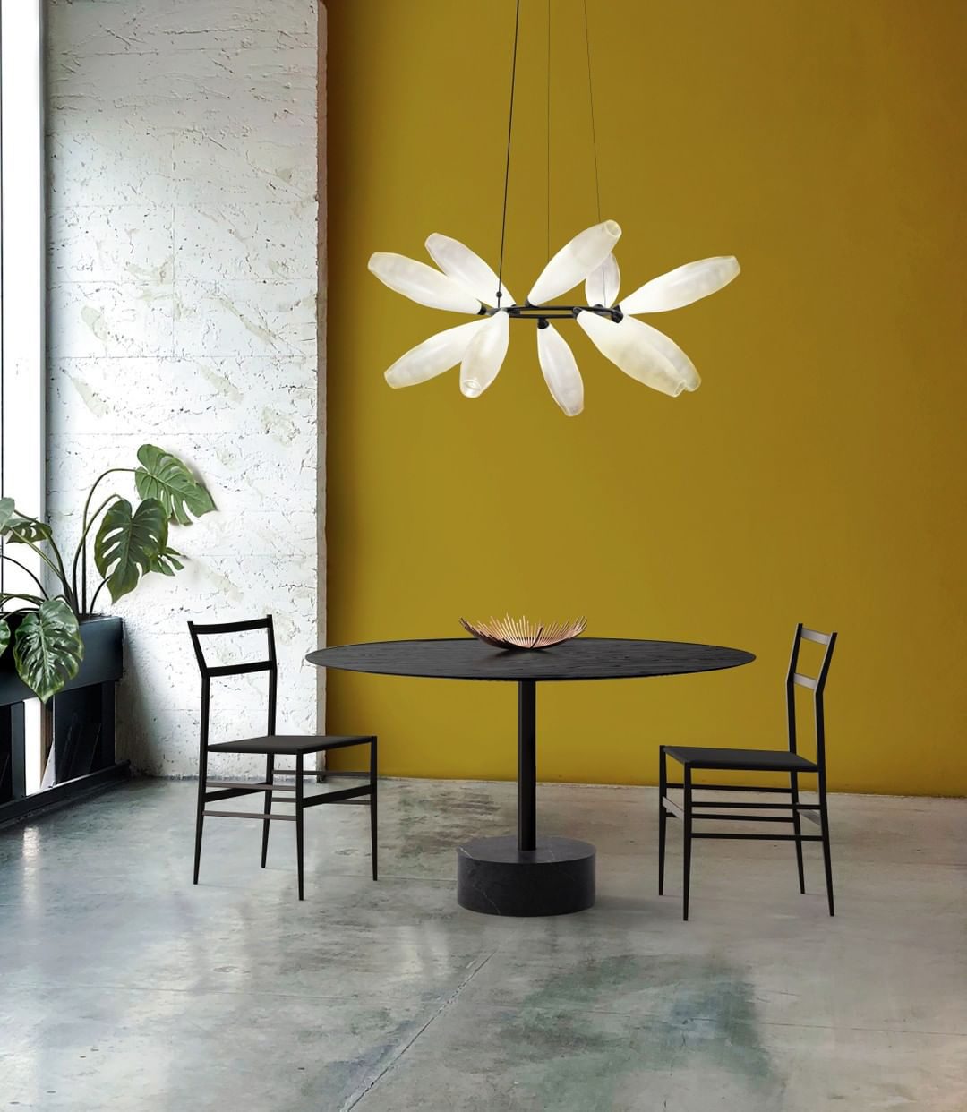 A round Clérachic chandelier of white leaves hangs against a mustard yellow wall above a small black table and two slim chairs.
