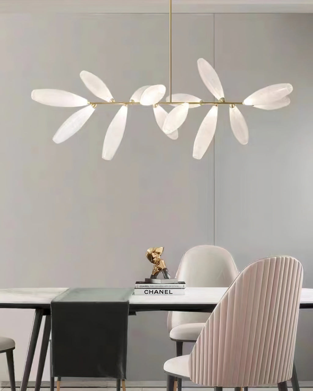 A linear Clérachic chandelier of frosted glass leaves hangs against a grey wall above a table with a black runner and a pink pleated chair.