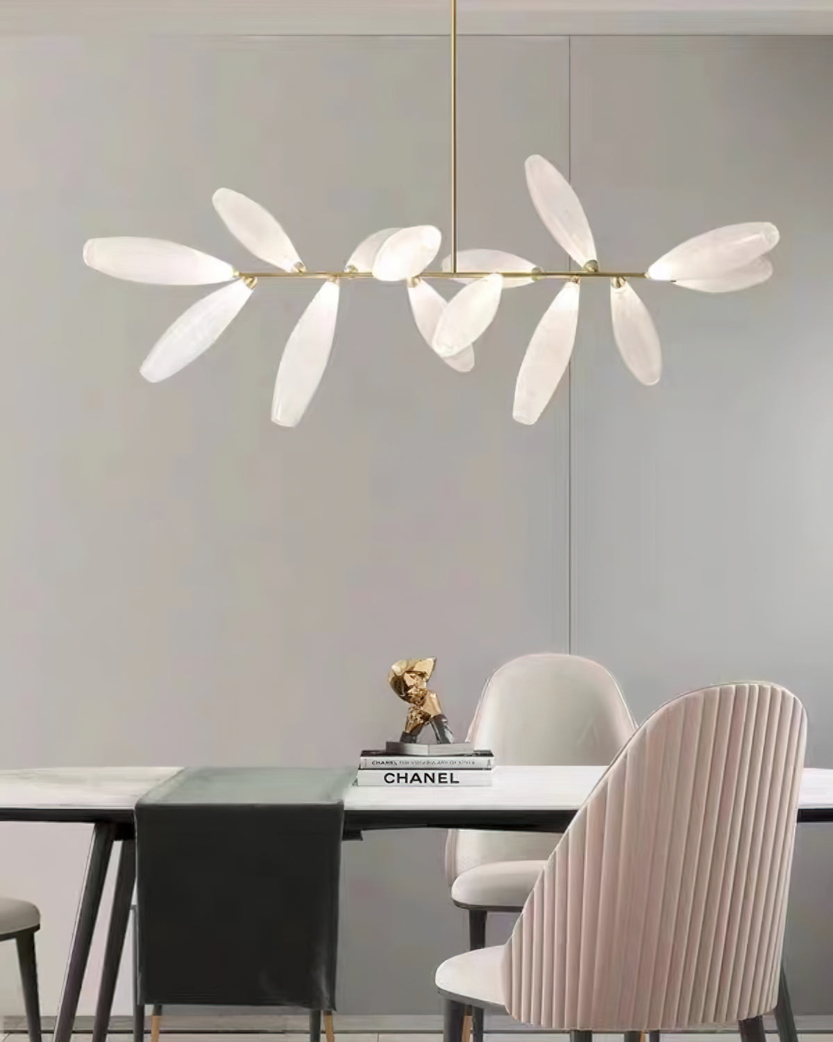 A linear Clérachic chandelier of frosted glass leaves hangs against a grey wall above a table with a black runner and a pink pleated chair.