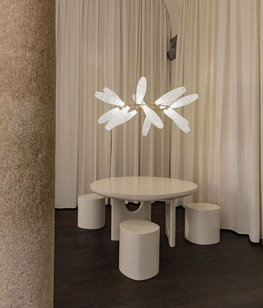 A small Clérachic chandelier of white glass leaves hangs above a round cream table and stools, in front of floor-length curtains.