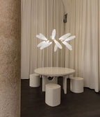 A small Clérachic chandelier of white glass leaves hangs above a round cream table and stools, in front of floor-length curtains.