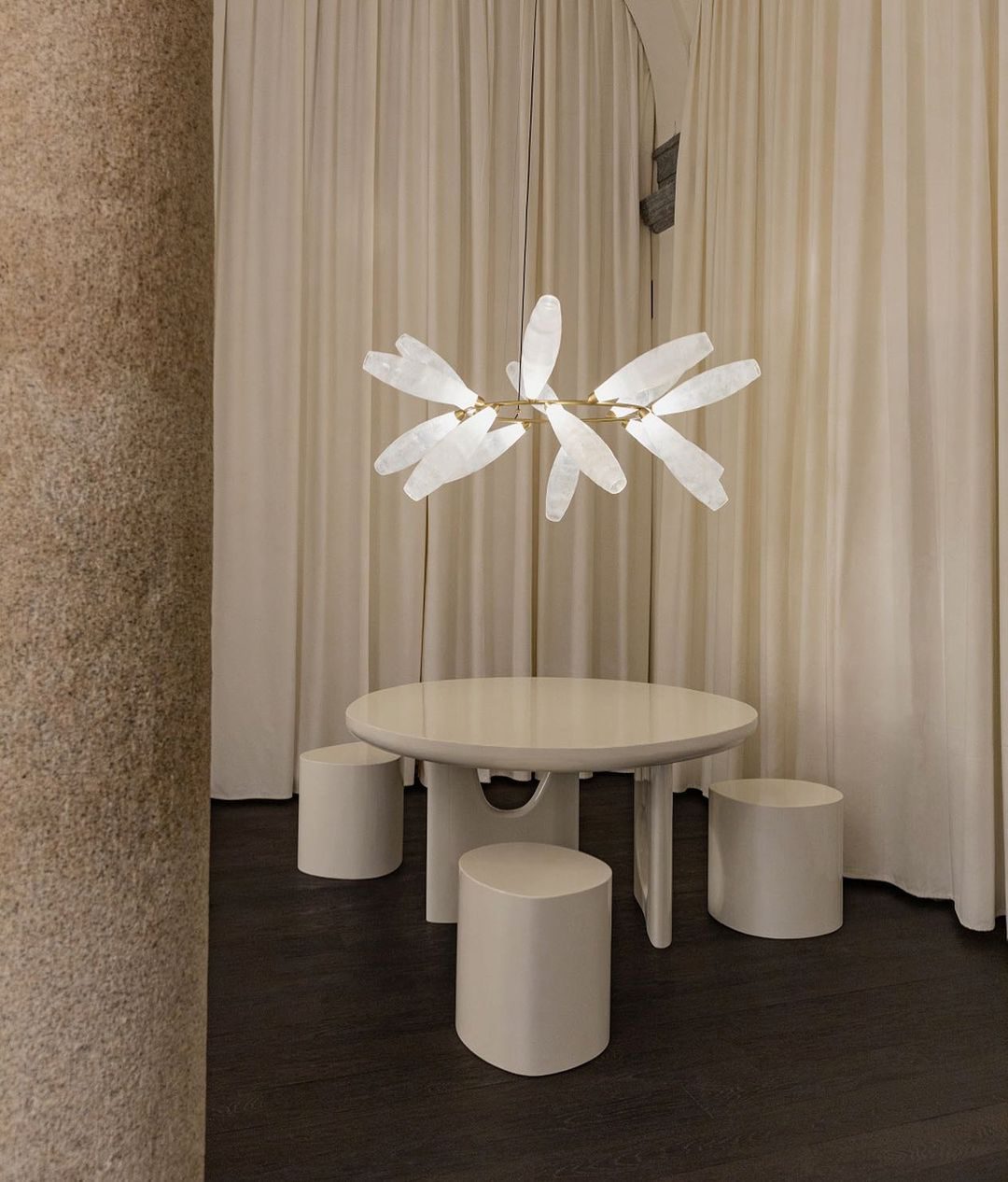 A small Clérachic chandelier of white glass leaves hangs above a round cream table and stools, in front of floor-length curtains.