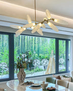A Clérachic chandelier of white glass leaves glows beneath a coved ceiling, with a large window onto trees and an oval marble table below.