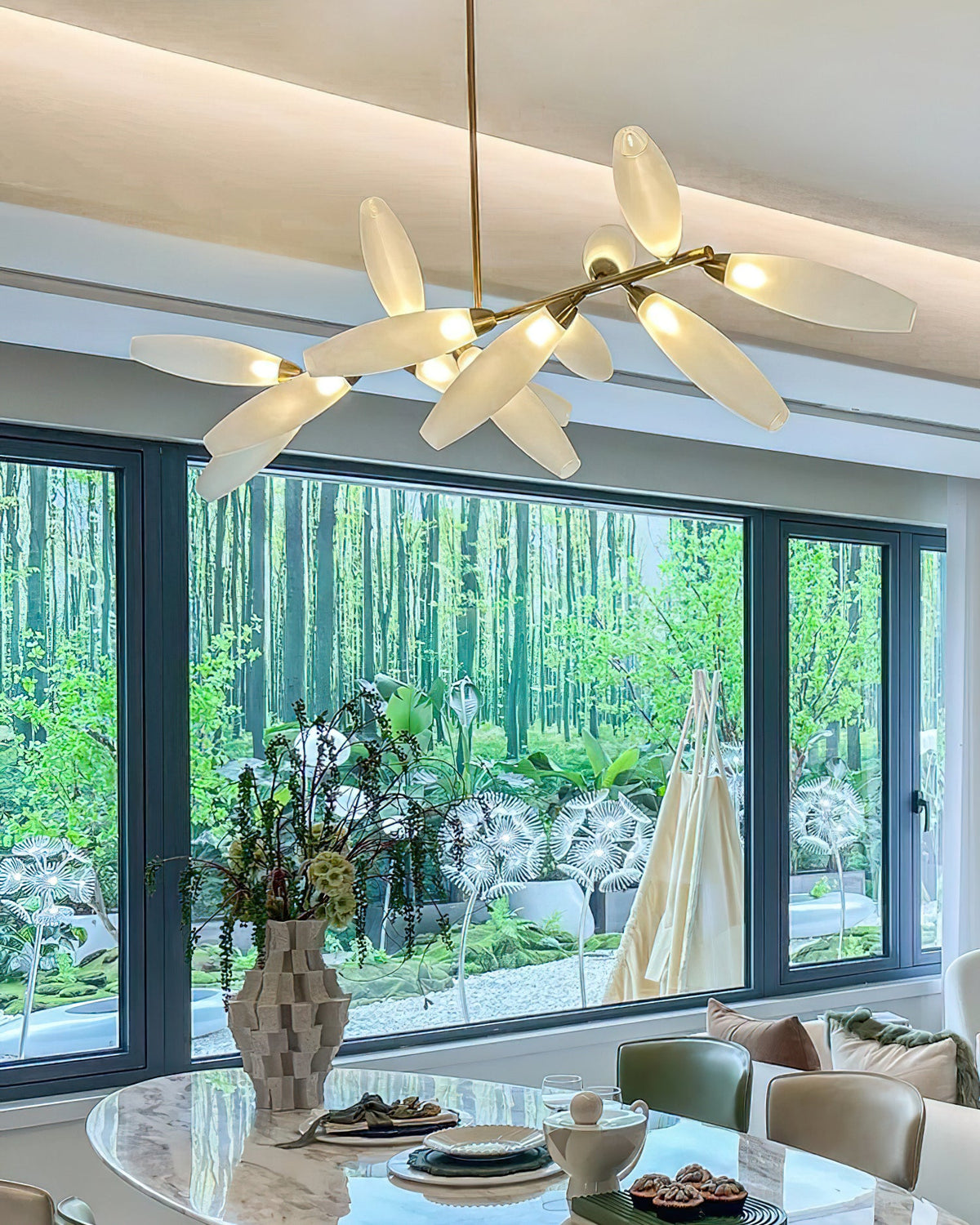A Clérachic chandelier of white glass leaves glows beneath a coved ceiling, with a large window onto trees and an oval marble table below.