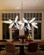A Clérachic chandelier of lit white glass leaves hangs above a round dark table with red flowers, in front of tall glazed doors.