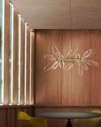 A Clérachic chandelier with clear glass leaves, a different finish from the white one, hangs on a wood-slatted wall above a dark round table.