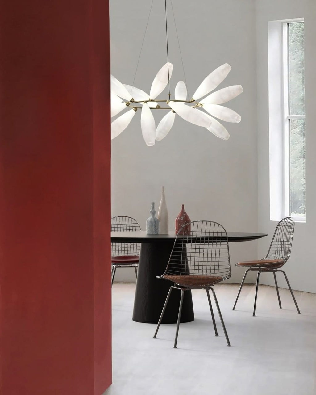 A round Clérachic chandelier of white leaves hangs beside a tall red wall above a black table with wire chairs and slim vases.