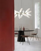 A round Clérachic chandelier of white leaves hangs beside a tall red wall above a black table with wire chairs and slim vases.