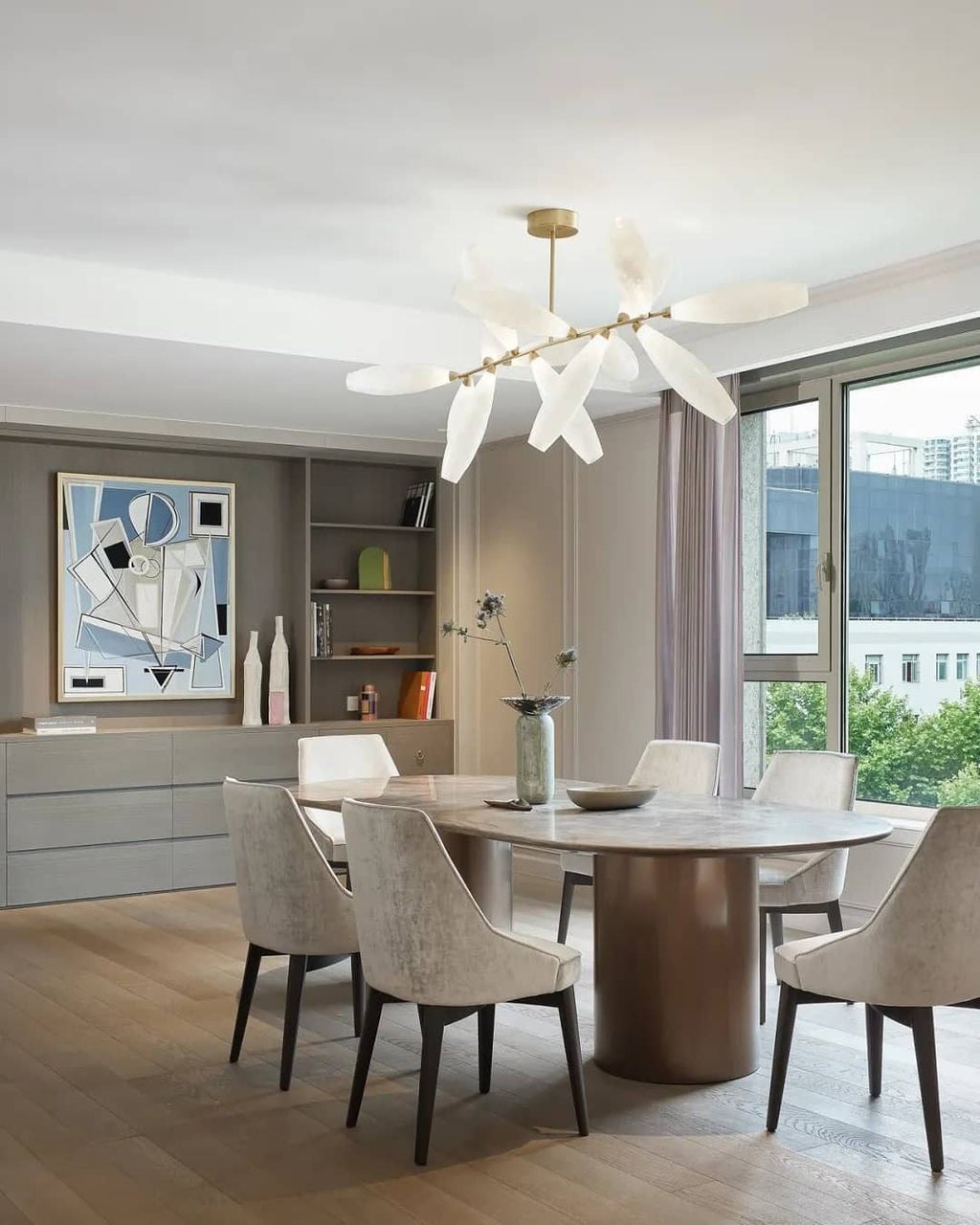A Clérachic chandelier of white glass leaves hangs from a gold canopy above an oval table with cream chairs, with a bright city window behind.