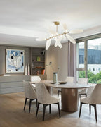 A Clérachic chandelier of white glass leaves hangs from a gold canopy above an oval table with cream chairs, with a bright city window behind.