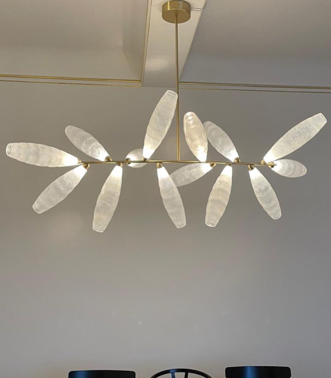 Close view of a linear Clérachic chandelier, its frosted white glass leaves on a gold rod against a grey wall.