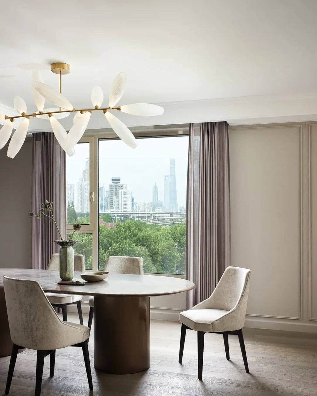The edge of a Clérachic chandelier of white glass leaves on a gold rod above a round table and taupe chairs, with a city view through the window.
