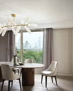 The edge of a Clérachic chandelier of white glass leaves on a gold rod above a round table and taupe chairs, with a city view through the window.