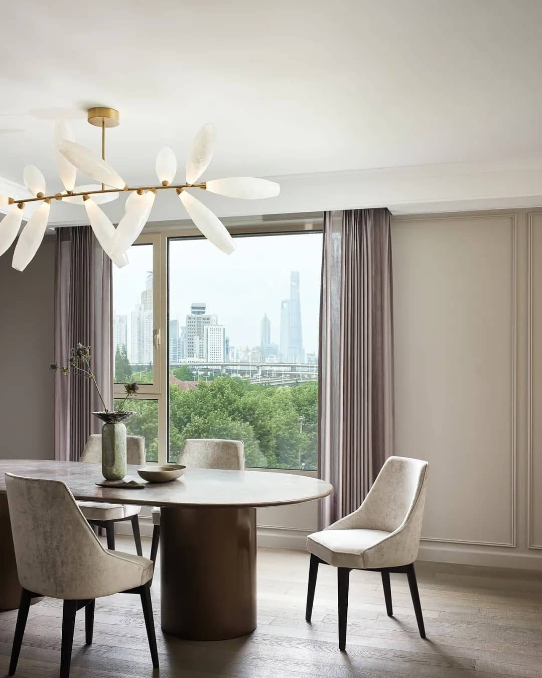 The edge of a Clérachic chandelier of white glass leaves on a gold rod above a round table and taupe chairs, with a city view through the window.