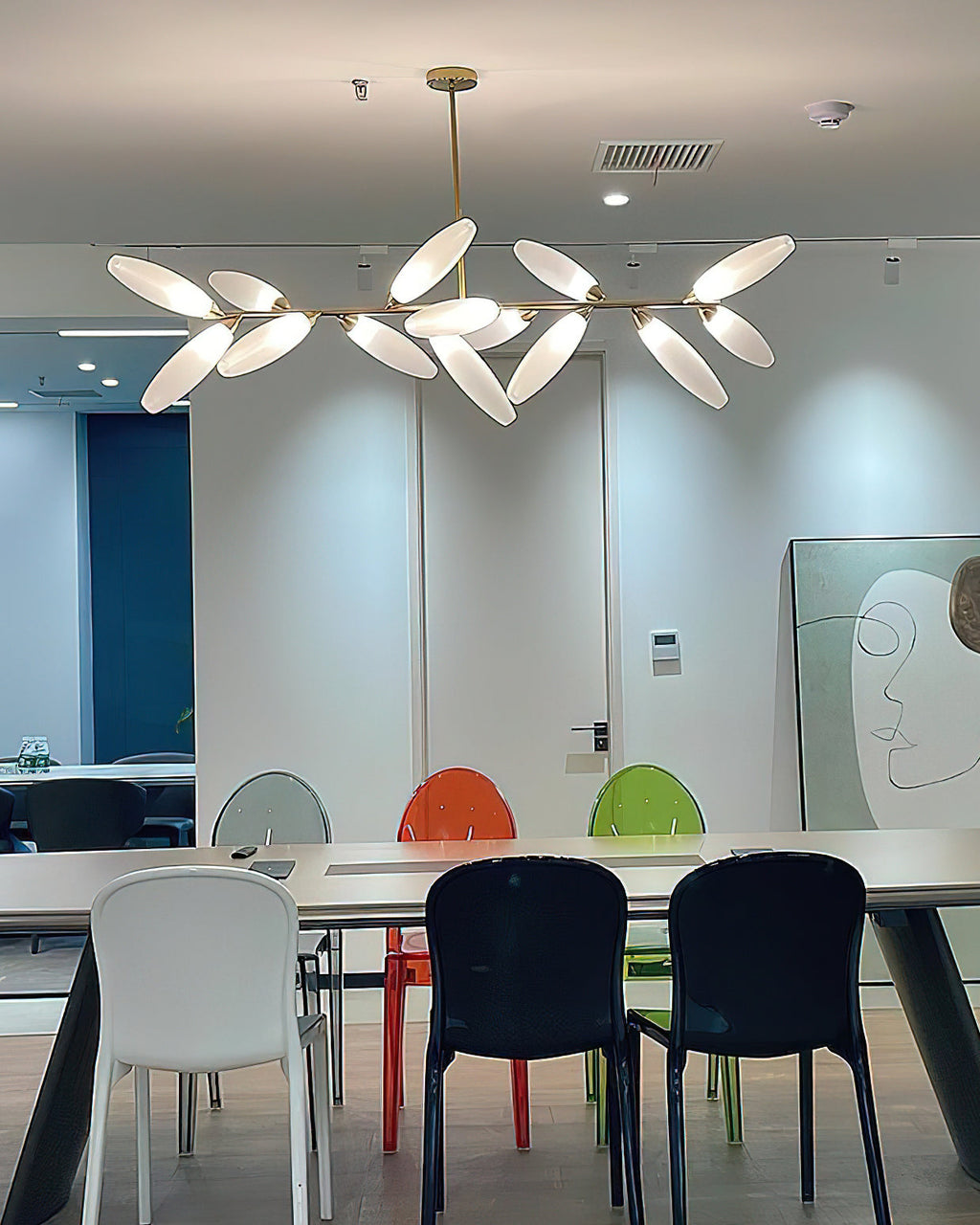 A linear Clérachic chandelier of lit white glass leaves hangs in a bright meeting room above a long table with white, black and coloured chairs.