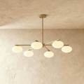 Clérachic six-head chandelier with slim brass arms and rounded opal glass shades, hung from a short stem in a plaster-walled room.