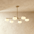 Clérachic six-head chandelier with slim brass arms and rounded opal glass shades, hung from a short stem in a plaster-walled room.