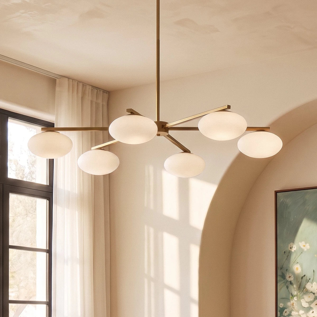 Close view of a Clérachic six-head chandelier, its oval opal shades glowing on brass arms beside a sheer curtain and arched wall.