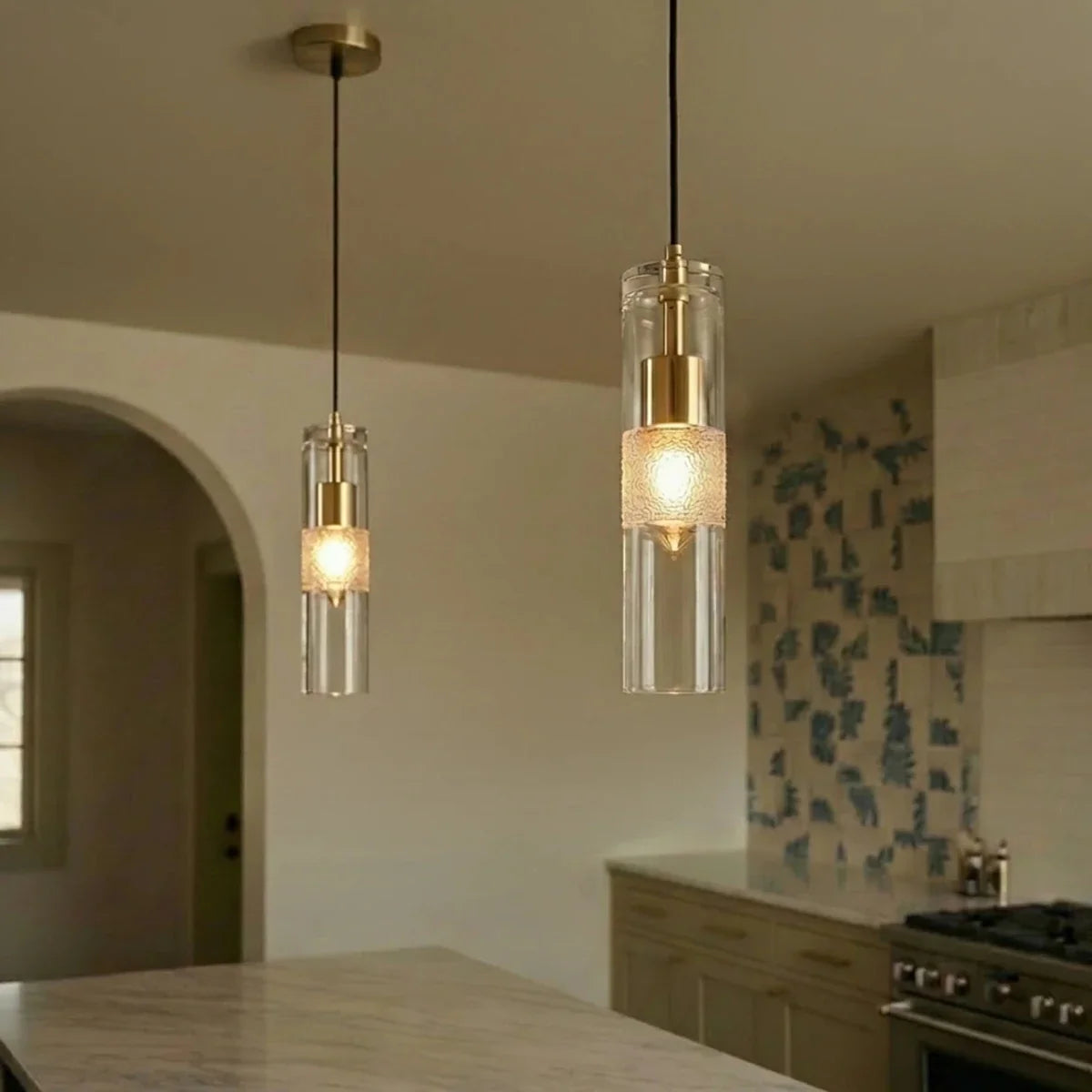 Clérachic glass cylinder pendants with brass holders and glowing frosted bands, hanging in a warm kitchen beside an arch.