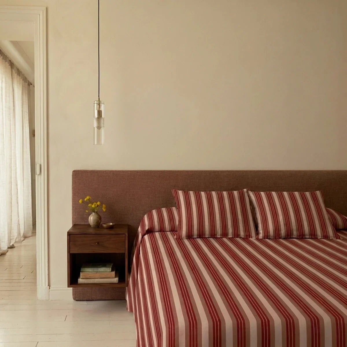 Clérachic slim glass cylinder pendant hung beside a bed with a floating walnut nightstand and red striped bedding.