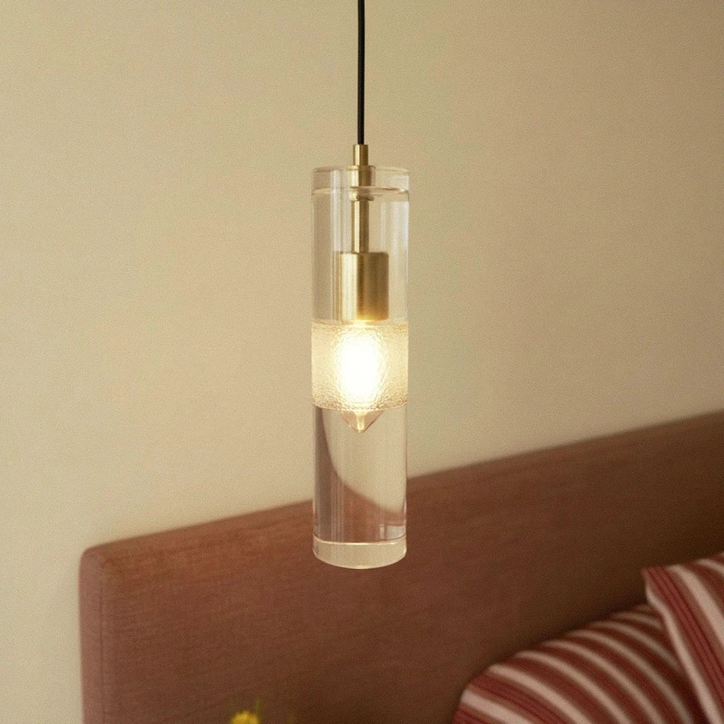 Clérachic lit glass cylinder pendant with a brass holder and frosted band, hanging by an upholstered headboard.