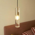Clérachic lit glass cylinder pendant with a brass holder and frosted band, hanging by an upholstered headboard.