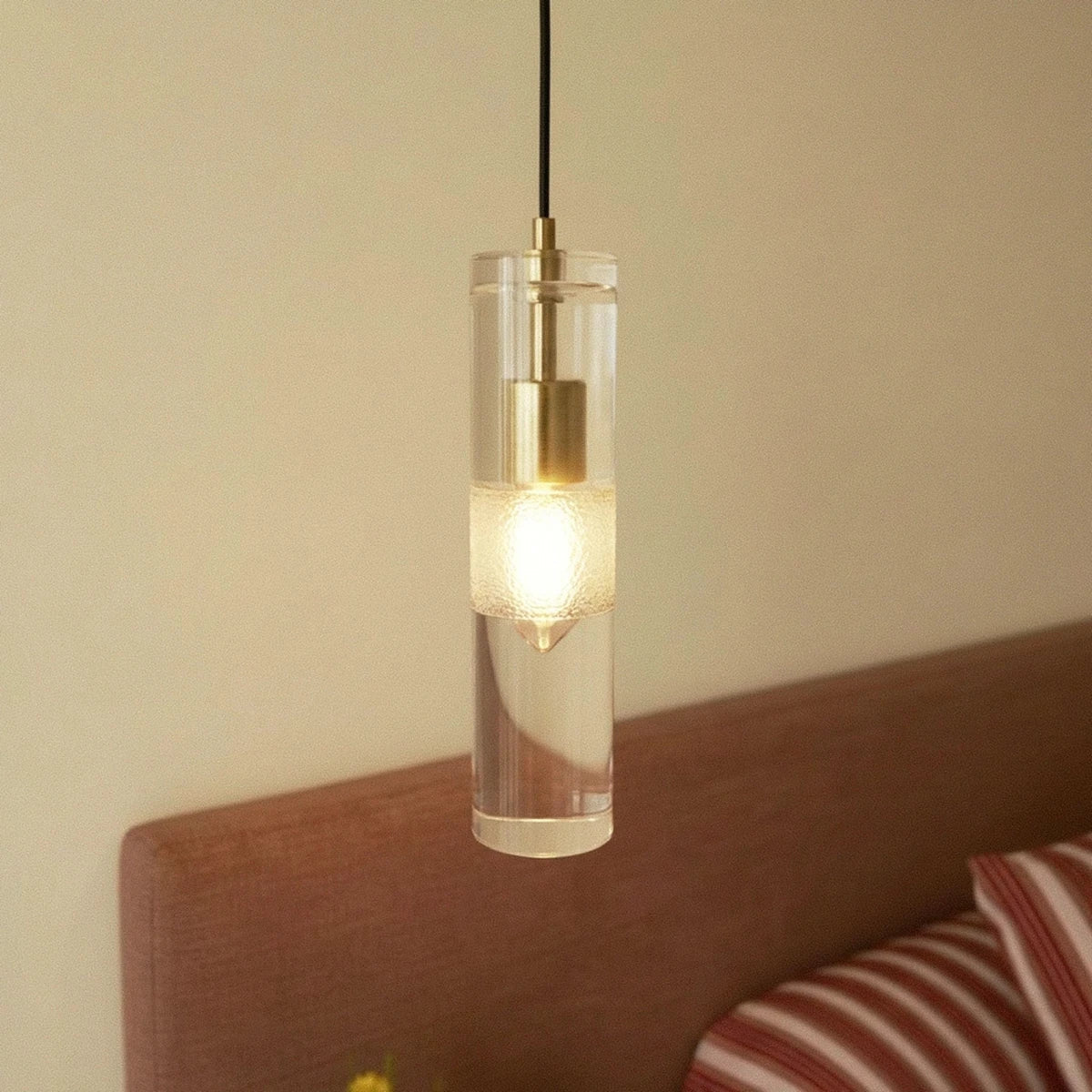 Clérachic lit glass cylinder pendant with a brass holder and frosted band, hanging by an upholstered headboard.