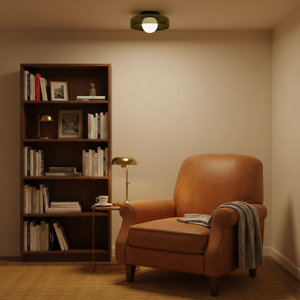 A Clérachic ceiling light with an olive glass disc hangs over a reading corner with a leather armchair, a bookcase and a brass lamp.