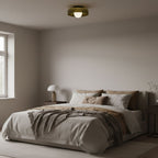 In a bedroom with a window and neutral bedding, a Clérachic glass-disc ceiling light sits above the bed with its globe lit.