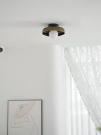A Clérachic dark glass-disc ceiling light with a white globe sits above a white room with lace curtains and framed line art.