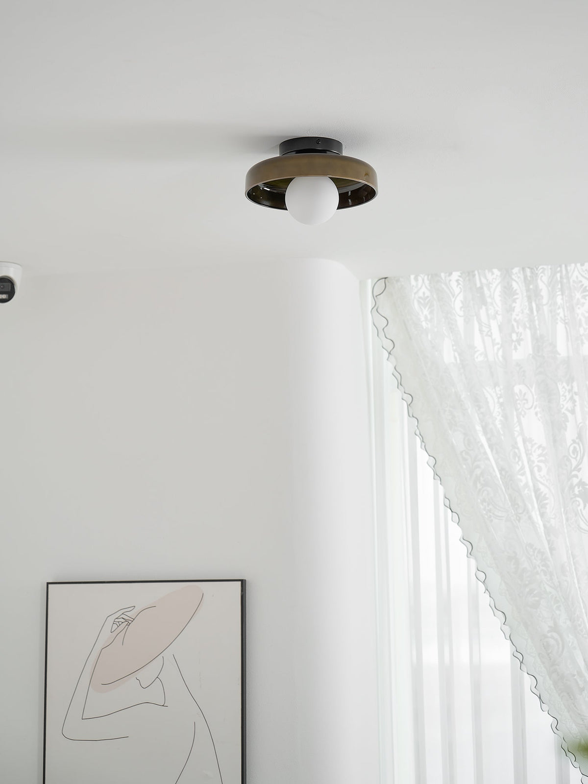 A Clérachic dark glass-disc ceiling light with a white globe sits above a white room with lace curtains and framed line art.