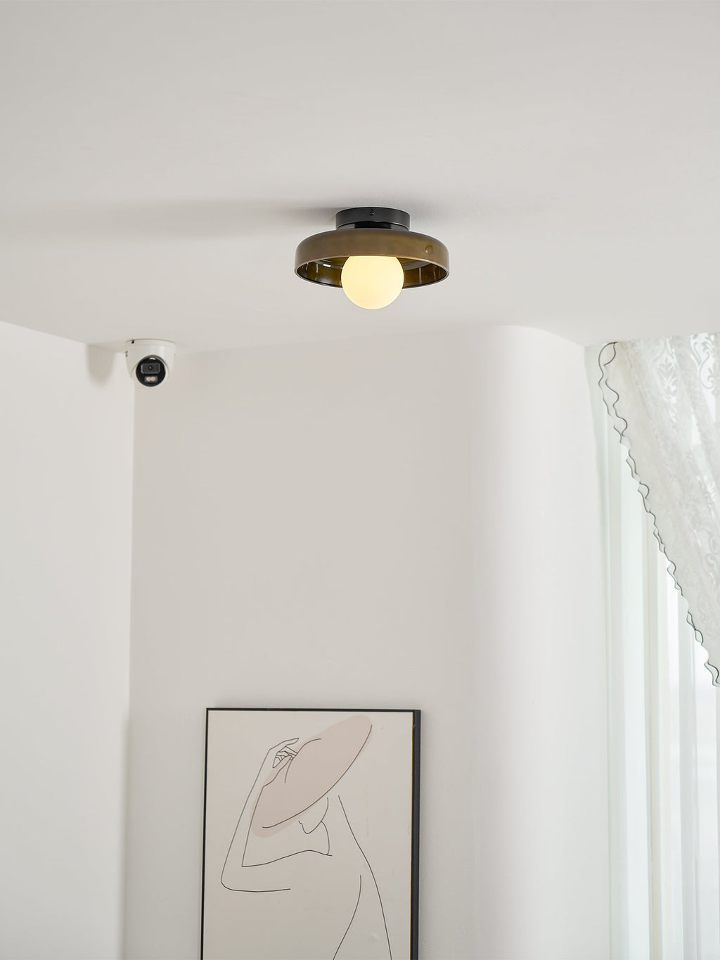 A Clérachic dark glass-disc ceiling light above a pale corner with a lace curtain and a line-drawing print.