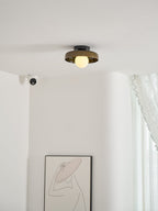 A Clérachic dark glass-disc ceiling light above a pale corner with a lace curtain and a line-drawing print.