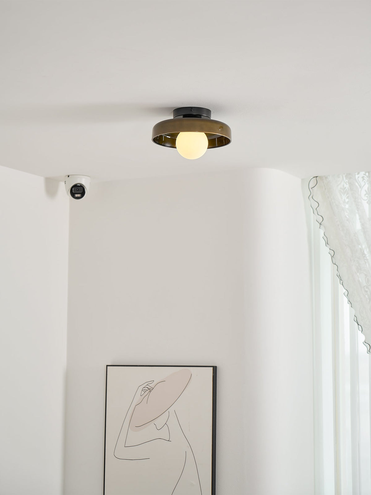 A Clérachic dark glass-disc ceiling light above a pale corner with a lace curtain and a line-drawing print.