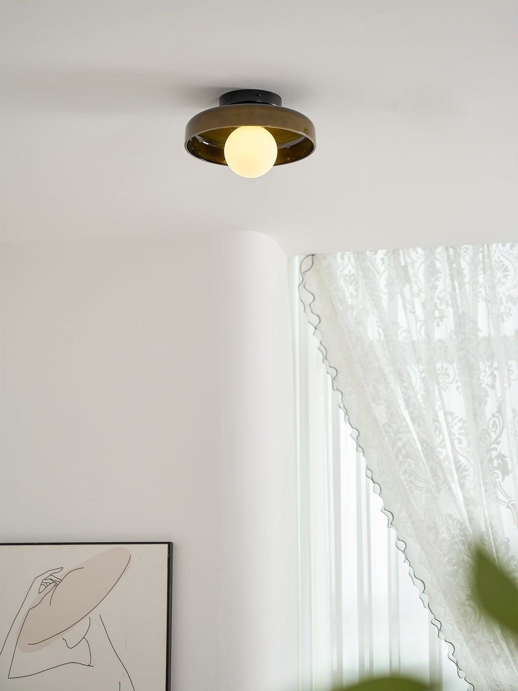 A Clérachic dark glass-disc ceiling light with a lit globe, above a curved white wall and a lace curtain.