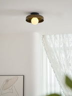 A Clérachic dark glass-disc ceiling light with a lit globe, above a curved white wall and a lace curtain.