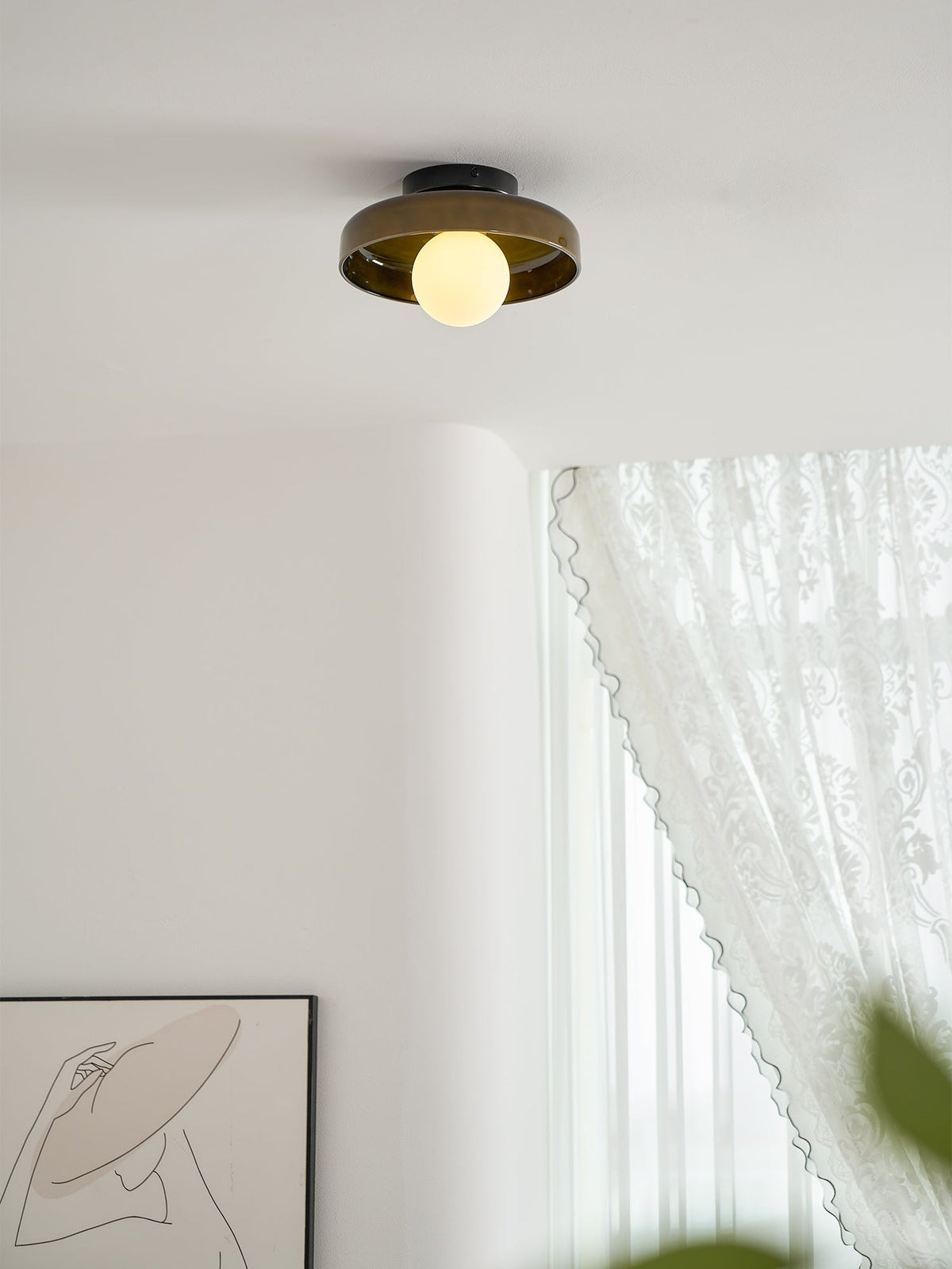 A Clérachic dark glass-disc ceiling light with a lit globe, above a curved white wall and a lace curtain.
