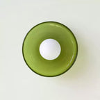 Head-on view of a clear green glass disc light from Clérachic with a white globe, on a pale green-white wall.