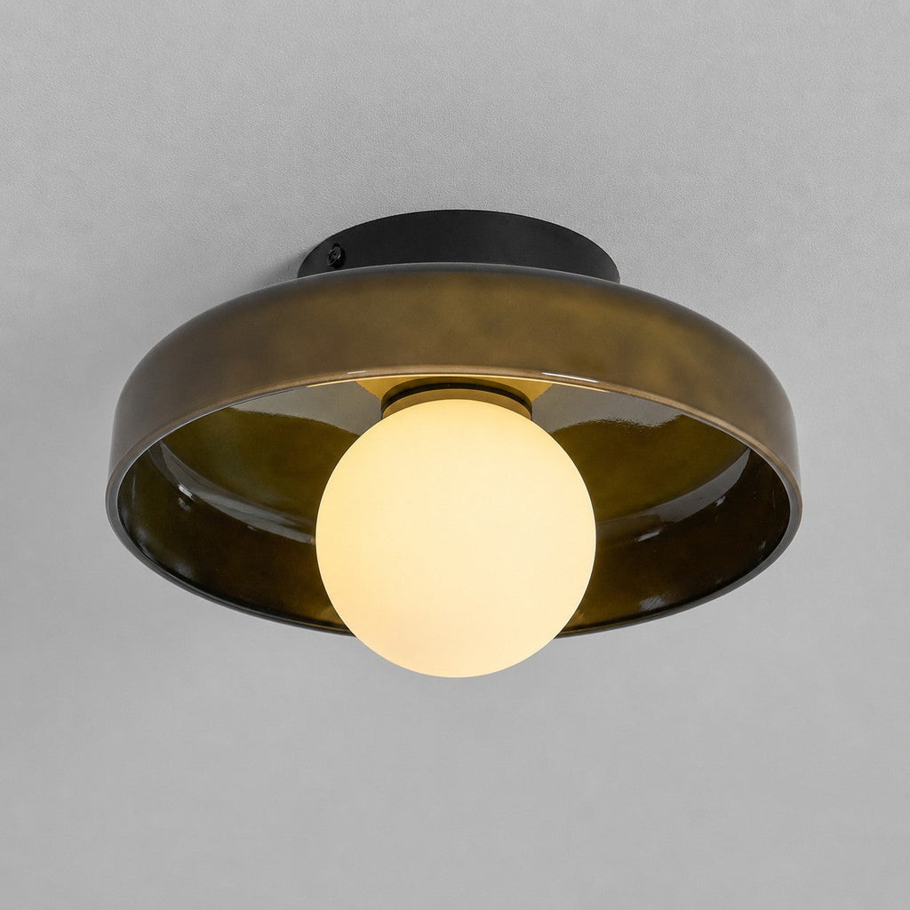 A Clérachic smoked olive-brown glass disc against a grey ceiling, with a black plate and its white globe lit warm.