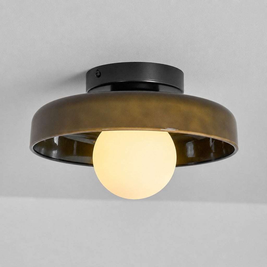 Side view of the Clérachic olive-brown glass-disc light, its white globe glowing warm under the rim against a grey ceiling.