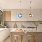 Two Clérachic glass pendants, one light blue and one amber, hang over a kitchen island with two pale wood stools.