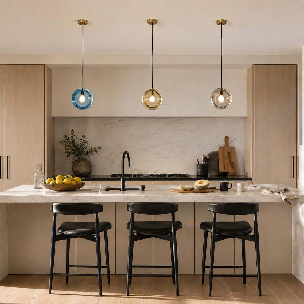 Three Clérachic glass pendants in light blue, amber and clear on brass ceiling caps, above a kitchen island with black stools.