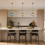 Three Clérachic glass pendants in light blue, amber and clear on brass ceiling caps, above a kitchen island with black stools.