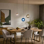 Clérachic glass pendants in pink, dark blue and amber hang at staggered heights over an oak dining table with grey chairs.
