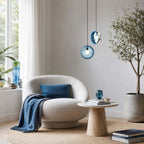 Two Clérachic glass pendants in light blue, beside a window and an olive tree, above a boucle armchair with a blue cushion.