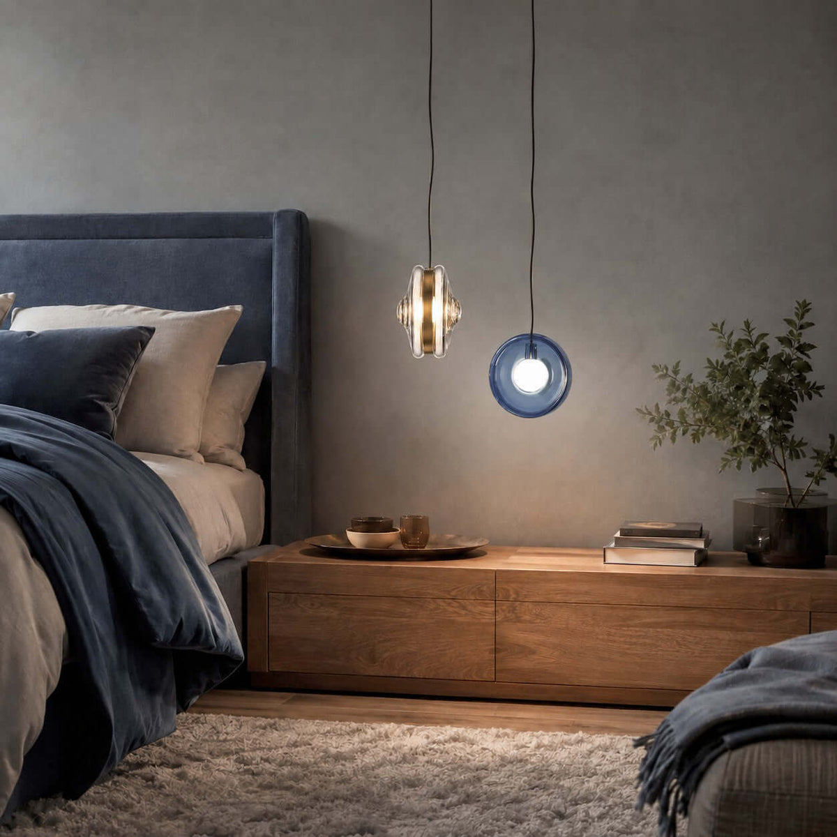 A clear and a dark blue Clérachic glass pendant hang beside a bed with navy linen, over a low wood chest in a grey bedroom.
