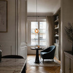 A single amber Clérachic glass pendant lit in a doorway, over a small round table and a dark armchair by a tall window.