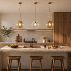 Three Clérachic glass pendants in amber, clear and amber, hung over a marble kitchen island with three curved wooden stools.