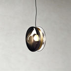 This Clérachic glass disc pendant in smoky grey glass, with a brass band and a clear rim, against a grey plaster wall.